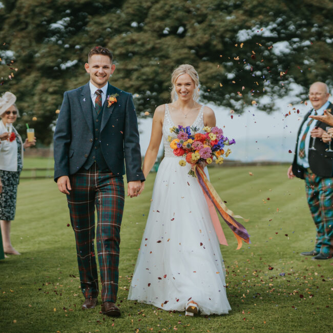 Newly wed bride and groom, walking hand in hand through confetti at their September wedding. Bride is holding a beautifully vibrant and fun floral bridal bouquet.